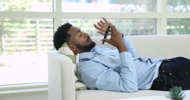 自宅でソファーに横たわっているスピーカーフォンにスマートフォンを抱えているケアフリーのアフリカ人男性は 友人と個人的な話し合いをリードし ニュースを共有し メッセンジャーアプリを使用してボイスメッセージを残します — ストック動画
