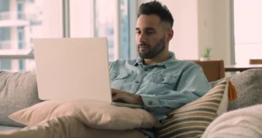 Ciddi yakışıklı İspanyol adam evde tek başına dizüstü bilgisayarda çalışıyor rahat bir kanepeye kurulmuş e-posta yazıyor, mesajları paylaşıyor, müşteriyle uzaktan iletişim kuruyor, işi uzaktan çözüyor gibi görünüyor.