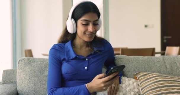坐在舒适的沙发上 坐在耳机里的女人 听流行的音乐 欣赏新的播客 与现代技术一起度过闲暇时光 品味高质量的声音 在线服务用户流 — 图库视频影像