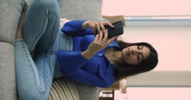 Genç Hintli kadın oturma odasındaki koltukta oturan cep telefonunu dikey olarak kullanıyor. Bekar tatmin olmuş kadın yeni cep telefonu e-randevu uygulamasından hoşlanıyor, mesajları paylaşıyor, sosyal medya ağlarındaki arkadaşlarıyla sohbet ediyor