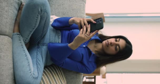 年轻的印度女人坐在客厅的沙发上用手机 纵观全景 心满意足的女性在社交媒体网络上享受新的移动电子约会应用 分享信息 与朋友聊天 — 图库视频影像