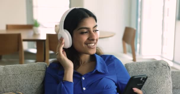 クローズアップケアフリーのインドの女性は 現代のワイヤレスヘッドフォンを介して音楽を聞きます 携帯電話を使用しています デジタルストリーミングオンラインサービス お気に入りの曲を使用してプレイリストをお楽しみください ソファで余暇を過ごす — ストック動画