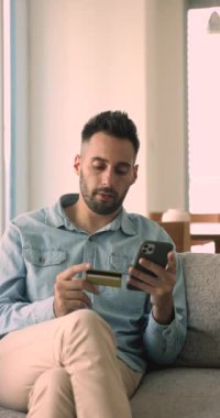 Dikey görünümlü yakışıklı 30 'lu yaşlarda İspanyol bir adam kart ve telefon tutuyor modern teknolojiyi kullanarak evden elektronik alışveriş yapıyor, kolay mobil uygulamalara para aktarıyor, internete para harcıyor.