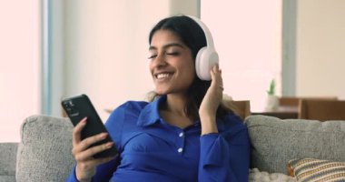 Büyük kulaklıklı mutlu, huzurlu, genç Hint müzik hayranı kız çevrimiçi, kapalı gözlerle en sevdiği melodileri dinliyor, gülümsüyor, ortamı rahatlatan şarkılardan hoşlanıyor, akıllı telefon tutuyor.