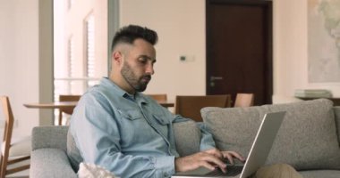 Ciddi Hispanik bir adam dizüstü bilgisayarla kanepeye oturur, kablosuz bilgisayarı kucağına alır, blog yazılarını düşünür, laptop ve internet bağlantılarını kullanarak evden internet üzerinden çalışır. Telework, iş için modern teknoloji