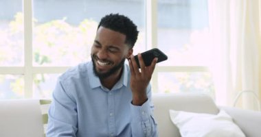 Genç Afrikalı adam evdeki koltukta oturuyor cep telefonunu dinliyor arkadaşından sesli mesaj alıyor, cep telefonu uygulamasını kullanarak cevap veriyor, sesli mesaj gönderiyor, gülümsüyor, uzaktan iletişimin keyfini çıkarıyor, modern teknolojinin tadını çıkarıyor.