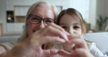Yaşlı büyükanne ve anaokulu torununun mutlu yüzlerini kapatın. Birleştirilmiş parmaklarıyla, sevgi ve bağlılık sembolü göstererek görülüyor. Çok nesilli aile anlayışı ve sevgisi