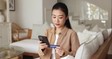 Akıllı telefon kartı kullanan Asyalı kadın evde e-alışveriş yapıyor, elverişli ve güvenli alışveriş deneyiminin tadını çıkarıyor, kolay bir ödeme ve alışveriş yapıyor, başarılı bir satın alma ile tatmin oluyor.