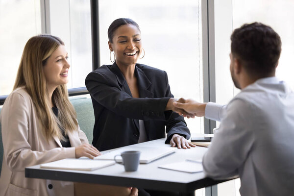 Happy multiethnic business male and female partners shaking hands over table, buying and selling startup, celebrating hiring, employment, partnership, investment, giving handshake