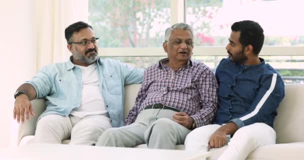 インドの多世代家族 ハンサムな若い25代男性は 成熟した50代の父親と70代の孫と時間をソファに座って 快適でフレンドリーな会話をリードし 笑顔は家で週末のレジャーを楽しんでいます — ストック動画