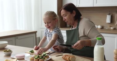 Evcil önlük giyen, tatlı küçük torun kızına yemek pişirmeyi öğreten taze sebzeli sandviç hazırlayan, jambon dilimlemeye yardım eden bir büyükanne. Büyükanne ve çocuk aşçılık hobisinin keyfini çıkarıyorlar.