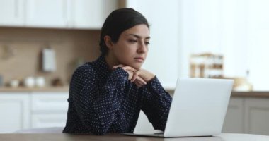 Düşünceli genç Hintli bağımsız kadın mutfak masasında dizüstü bilgisayar kullanıyor, iş yerinde çalışıyor, bilgisayardaki internet projesini düşünüyor, daktilo ediyor, karar veriyor, ekrana bakıyor.