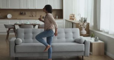 Mutlu aktif genç Hintli kadın evde dans ediyor, gri stil ev kanepesinde hoplayıp zıplıyor, eğleniyor, eğleniyor, eğleniyor, eğleniyor, müzik, hareket ediyor, yeni rahat bir dairede oturuyor.