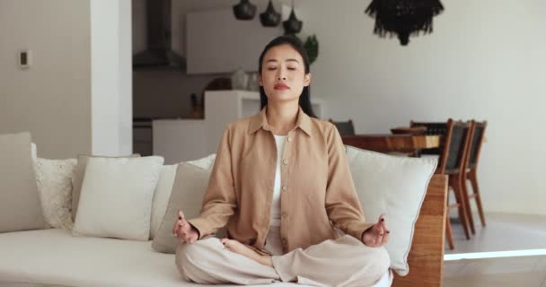 女性瞑想は目を閉じて蓮の位置にソファーに座ります 女性は心を落ち着かせることによってストレスを減らし 感情的な幸福を高め 毎日のルーチンを実行し 健康的な生活習慣を保ちます スピリチュアルな成長 — ストック動画