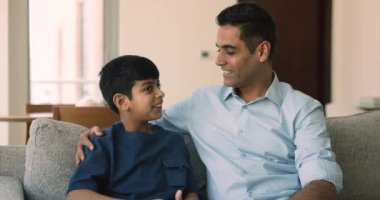 Gülümseyen orta yaşlı Hintli bir adam küçük 8 'li yaşlardaki oğluna sarılıyor. Güvenilir bir konuşma yapıyor. Evdeki kanepede birlikte oturuyorlar. İletişim, bakım ve güven, ebeveynler ve çocuklar arasındaki anlayış