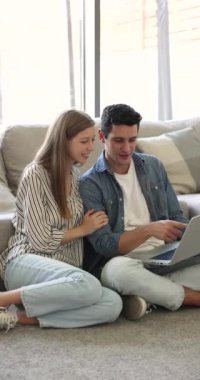 Oturma odasında genç İspanyol çift rahat bir şekilde dizüstü bilgisayarlarıyla modern e-ticaretle ilgileniyor, ürünleri ve hizmetleri değerlendiriyor, hafta sonu gezisi için bilet alıyor, evleri için eşyalar seçiyor.