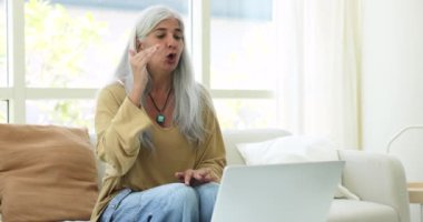 Olgun bir kadın video konferansında otururken kanepede oturup laptopa bakar, konuşma ve açıklamalar yapar, çevrimiçi sınıfa öncülük eder, müşterilerine profesyonel danışmanlık hizmeti verir, uzaktan çalışırlar.