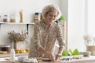 Gülümseyen olgun bir kadın kurabiye yapıyor, ev yapımı tatlı hazırlıyor, hamuru şekillendiriyor, hafta sonları hamur işi yapıyor, yemek masasında mutfakta duruyor, aşçılığın tadını çıkarıyor. Hoş ev işleri, hobi, aşçılık.