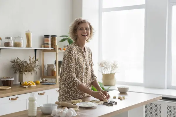 ポジティブな中年主婦を笑い 現代のキッチンで料理し カメラを見つめ 自然の生態学的な製品とテーブルに立って 生地をひざまずき パンを作る 自宅で週末にクッキーを作る ロイヤリティフリーのストック画像