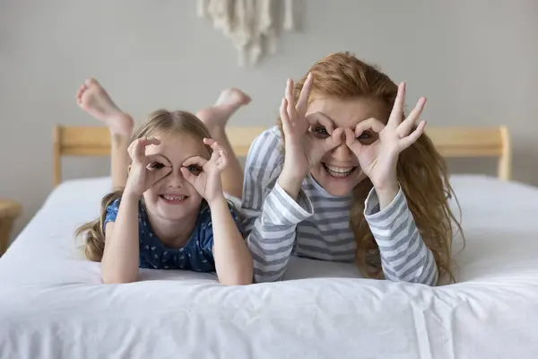幸せな小さな遊び心のある娘と若い母親は 指を通してカメラを見てベッドに横たわり 双眼鏡 またはアイウェアの形を作ります 楽しんでいる家族 広告ビジョンチェック 視力ケア ストックフォト