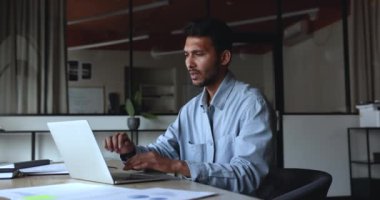 Ciddi anlamda Hintli genç proje lideri dizüstü bilgisayarda çalışıyor, işyerinde oturuyor, daktilo ediyor, başka yere bakıyor, çeneye dokunuyor, başlangıç için yaratıcı fikirler düşünüyor, gülümsüyor, başını sallıyor.