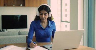 Kulaklıklı, konsantre olmuş genç bir kadın oturma odasında oturup online derste notlar yazarak yeni bilgiler ediniyor. Kız öğrenci çevrimiçi öğretmenin yardımıyla egzersiz yapıyor, dil öğreniyor, webinar izliyor