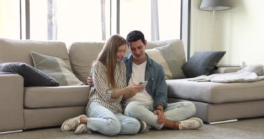 Gülümseyen İspanyol aşığı çift, modern akıllı telefonuyla ferah oturma odasında dinleniyor, çevrimiçi iletişimin tadını çıkarıyor, sipariş veriyor, e-servisleri kullanıyor, internette vakit geçiriyor.