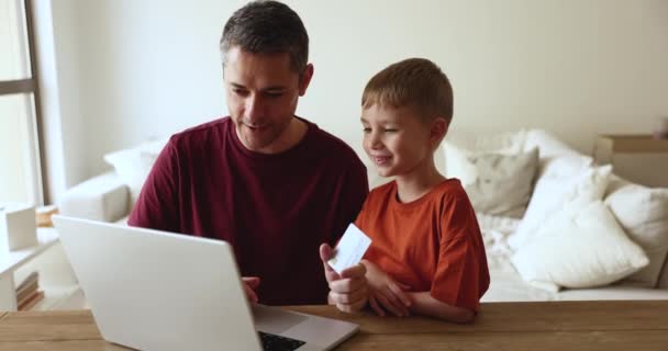 父と息子は デビットカードとラップトップを使用してオンラインショッピングを楽しんで 購入を議論し リビングルームのテーブルに座って支払いをする インターネットでギフトや電子サービスを購入する電子商取引のクライアント — ストック動画