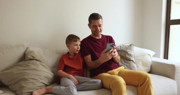 愛する父親は 小さな息子に新しいクールなモバイル面白いアプリケーションを示し スマートフォン画面を見つめるソファに座って 家族にビデオコールを楽しんで オンラインで楽しんで インターネットで自由な時間を過ごす — ストック動画