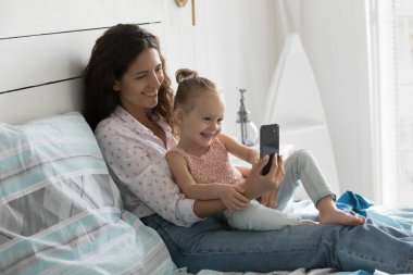 Genç anne, küçük bir çocukla yatakta oturup cep telefonunu tutarak ekrana bakarak, yeni uygulamalara bakarak, komik videolar izleyerek, annesini seven, eğitici online program programlarını izleyerek zaman geçiriyor. Ebeveyn kontrolü, modern teknoloji.
