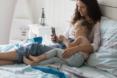 Genç, sevgi dolu anne yatakta dinleniyor, küçük tatlı kızına sarılıyor, aile hafta sonunu evde birlikte geçiriyor, video izlemek için cep telefonunu kullanıyor, internetten alışveriş yapıyor, yeni uygulamaların tadını çıkarıyor. İnternette boş zaman