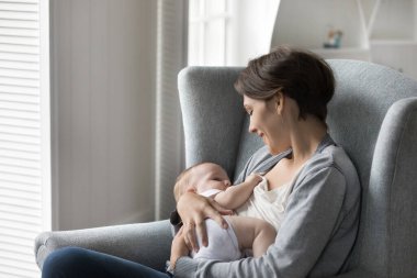 Genç ve sevecen anne, yeni doğmuş sevimli bebeği emzirmek için evdeki rahat koltuğa oturur. Eksiklik, büyüme için beslenme kaynağı ve çocukların iyi gelişimi, mutlu annelik anları, annelik.