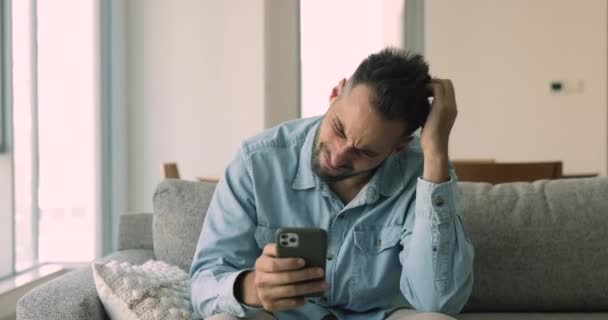 怒っているヒスパニックの男がソファーに座ってスマートフォンを見つめ メッセージの悪いニュース 壊れたデバイス 無線Lan接続が失われた ガジェットの問題 アプリのクラッシュに不満を感じる — ストック動画