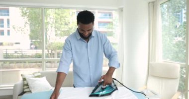 Modern oturma odasında tek başına duran, beyaz banyo havlularını ütüleyen, gülümseyen, günlük ev işlerini yapan, gündelik işlerinde yardımcı olan, mavi gömlekli yakışıklı yalnız bir Afrikalı. Oda servisi.
