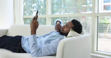 Tatmin olmuş Afrikalı bekar adam akıllı telefonu evdeki kanepede uzanıyor, mesajları paylaşıyor, internetin keyfini çıkarıyor, yeni mobil uygulama kullanıyor, sipariş veriyor, video seyrediyor. Modern teknoloji, yaşam tarzı, alışkanlık