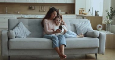 Güzel Latin bir kadın küçük sevimli kızına sarılıyor komik cep telefonu uygulamasını gösteriyor, selfie fotoğrafları çekiyor, online içerik videoları izliyor, evdeki koltukta birlikte gülüyor modern cep telefonu cihazıyla.