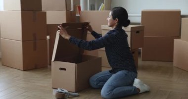 Pozitif genç Hintli ev sahibi kadın, taşınmak için karton kutuyu ayarlıyor, arka planda istiflenmiş kağıt kaplarla yerde oturuyor, yeni daireye taşınmak için eşyaları paketliyor,