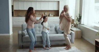 Neşeli enerjik üç kadın nesiller dans ediyor modern oturma odasındaki sıcak ahşap zeminde yalınayak zıplıyorlar, müzik dinliyorlar, tasasız görünüyorlar, hafta sonları eğleniyorlar, aktif aile evde eğleniyor.