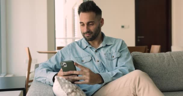30多岁的西班牙男人坐在沙发上 拿着手机 选择网上购物服务 在社交媒体上聊天 享受电子约会服务 使用新的移动应用程序寻开心 在家里消磨时光 — 图库视频影像