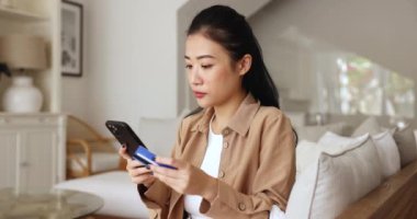 Asyalı bir kadın e-banka aracılığıyla kredi kartı ve telefon kullanarak ödeme yapmaya çalışıyor, stresli hissediyor, teknik sorunlar yaşıyor, çevrimiçi işlemler ve potansiyel dolandırıcılık konusunda endişeli, şüphe uyandırma girişimlerinde bulunuyor