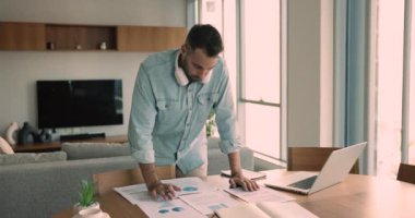 Genç Hispanik girişimci evde uzaktan çalışıyor, finansal verileri analiz ediyor, kağıt grafiklerini inceliyor, bilgiyi onaylıyor, notlar alıyor, strateji geliştiriyor, son teslim tarihi evrakları hazırlıyor, modern teknoloji kullanıyor.