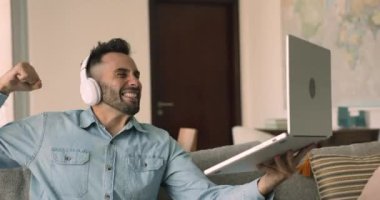 Genç mutlu İspanyol adam oturma odasında kanepede oturuyor kulaklık takıyor dizüstü bilgisayarında futbol maçı seyrediyor. Heyecanlı adam favori futbol takımının galibiyetini kutluyor. Dijital yayın hizmetleri, hobi