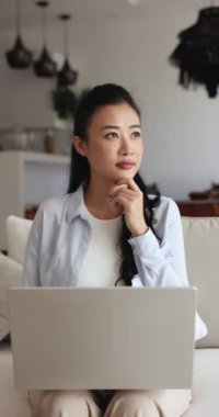 Ciddi bir kadın kanepeye oturur ve modern dizüstü bilgisayarla dikey olarak bakar. Uzaktan çalışma, hafta sonları sosyal medyada gezinme, çevrimiçi alışveriş, araştırma ve öğrenim istemcisi