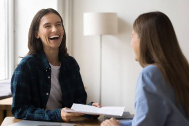 Neşeli proje yöneticisi kız iş arkadaşıyla günlük kıyafetlerle konuşuyor, gülüyor, eğleniyor, ofiste gayri resmi iletişimden keyif alıyor, iş belgelerini tutuyor, iş yerinde oturuyor.