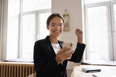 Neşeli, başarılı, cep telefonu tutan genç iş kadını, kazanan el hareketi yapıyor, mutlu hissediyor, iyi haberler hakkında seviniyor, başarıyı, başarıyı kutluyor, iş yerinde kazanıyor.