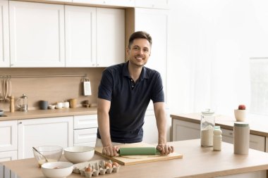 Neşeli, yakışıklı fırıncı evde yemek pişiriyor, mutfak masasında hamuru yuvarlıyor, kameraya bakıyor, portre için gülümsüyor. Mutlu genç adam ev yapımı hamur işi pişiriyor, aşçılıktan zevk alıyor, hazırlık yapıyor.