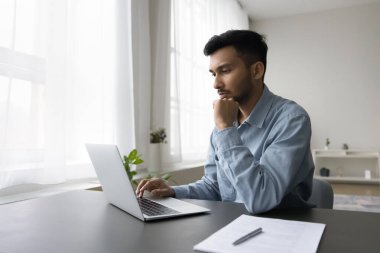 Ciddi meşgul genç Hintli serbest çalışan dizüstü bilgisayar başında oturuyor, ofis masasında oturuyor, bilgisayar başında çalışıyor, online uygulama kullanıyor, iş iletişimi için internet teknolojisi kullanıyor.