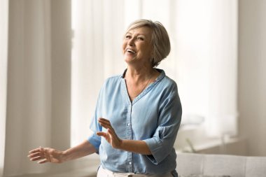 Heyecanlı, neşeli, olgun bir kadın evde eğleniyor, eğleniyor, eğleniyor, eğleniyor, eğleniyor, sağlıklı otlar ve kemikler modern dairede müzik eşliğinde dans ediyor, gülümsüyor, gülüyor.