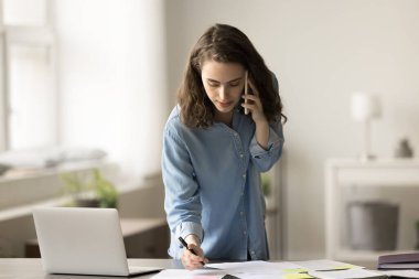 Ofis masasında cep telefonuyla konuşan meşgul genç Z iş kadını, kağıt raporlarını kontrol ediyor, notlar yazıyor, proje planını tartışıyor, cep telefonuyla başlangıç stratejisini tartışıyor.