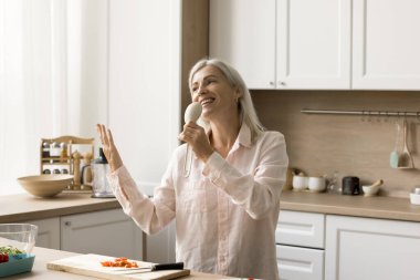 Olumlu heyecanlı olgun bir kadın mikrofon pişirmede şarkı söylüyor, malzemelerle masada dans ediyor, sebze dilimliyor, akşam yemeği için salata hazırlıyor, sağlıklı besleniyor, eğleniyor.